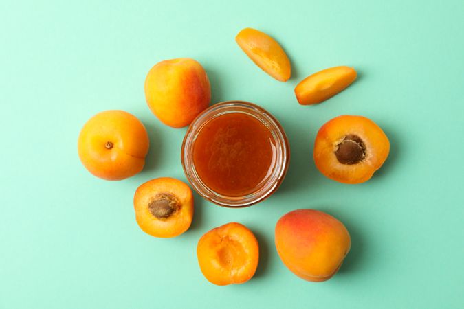 Glass jars with apricot jam and ingredients on mint background, top view