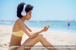 Woman in yellow bikini sitting on beach and looking at phone 0g2K3b