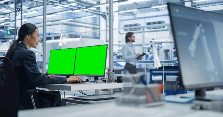 Female Robotics Technician Working on Desktop Computer with Green Screen Chromakey Mock Up Display in a Factory Office. Technological Research and Development Facility with Autonomous Assembly Line