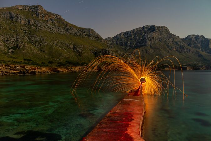 Person with burning circle on stones between water near hills at night