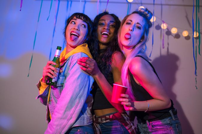 Three young women standing together making faces while holding drinks at a house party