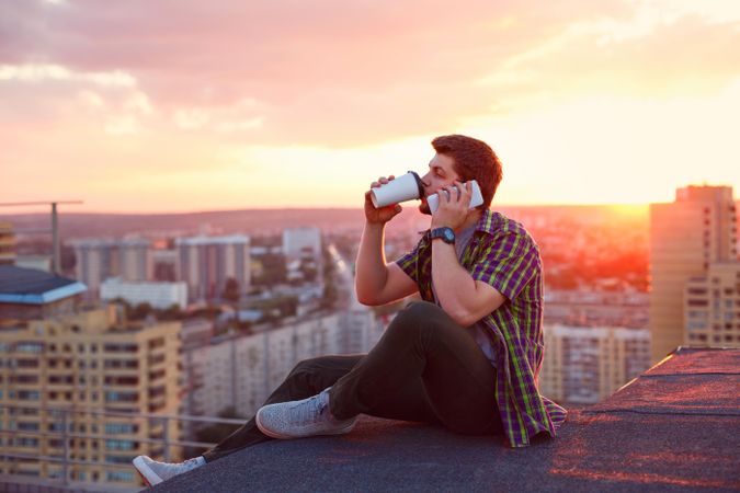 Male taking phone call while sitting on roof drinking takeaway coffee