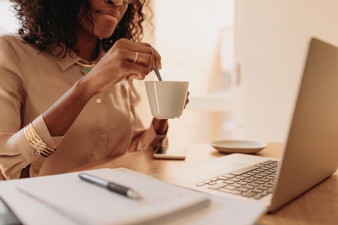 Businesswoman working on laptop computer stirring coffee at desk at home