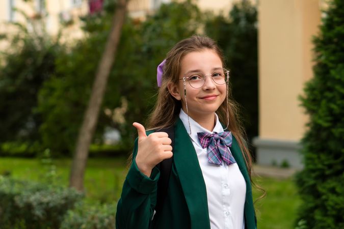 Young student with glasses giving thumbs-up outdoors.