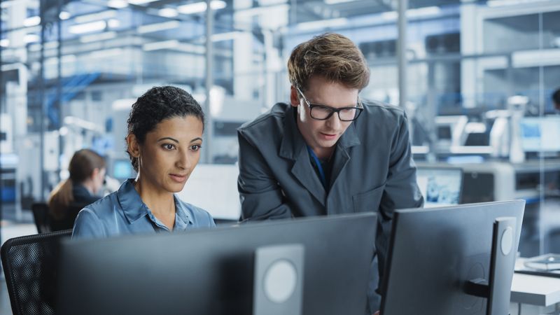 Portrait of a Young Male Assembly Line Worker and Hispanic Female Engineer Collaborating on a Project, Using Computer and Talking in a Factory with Robotic Arms Producing Modern High-Tech Devices.