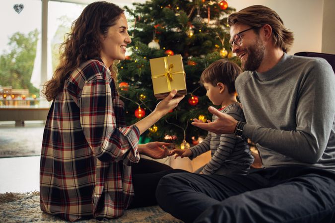 Couple sitting by Christmas tree with their son exchanging gifts at home