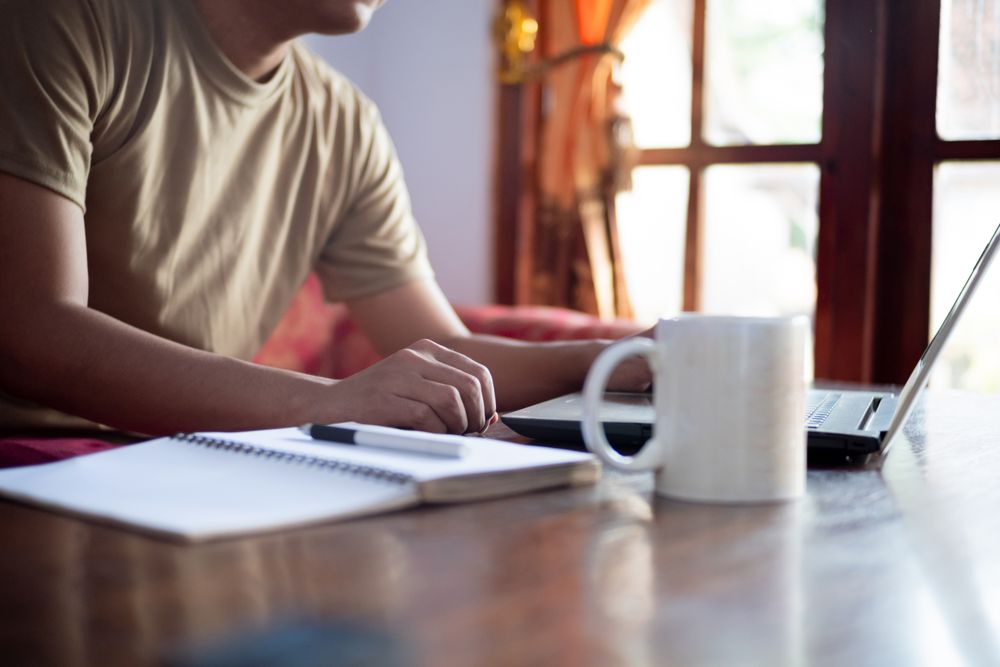 Asian Male with shirt working from home in living room with book, laptop and cup of coffee.