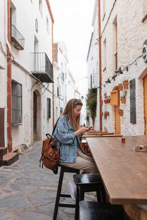 Side view of woman tourist in small village sitting at a table outside using a smartphone