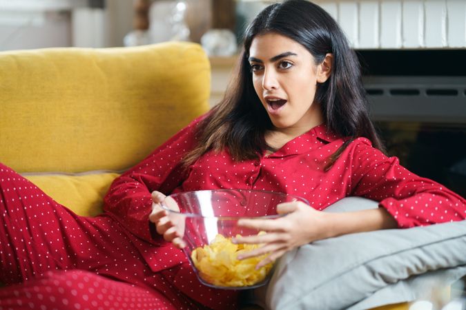 Woman in red pajamas relaxing on sofa and eating chips