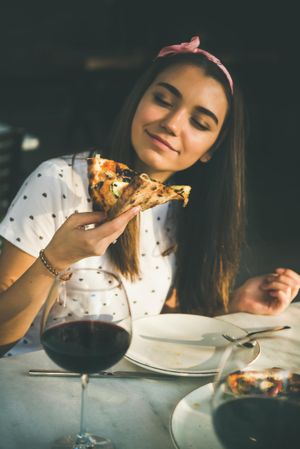Young woman eating pizza outside in the sun, with red wine