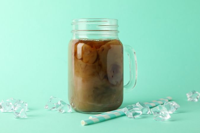 Jar of ice coffee, ice cubes and straws on mint background