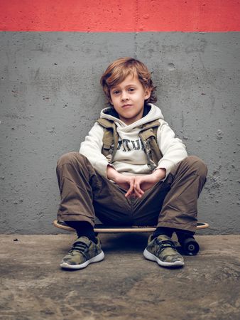 Cool child sitting on skateboard near wall
