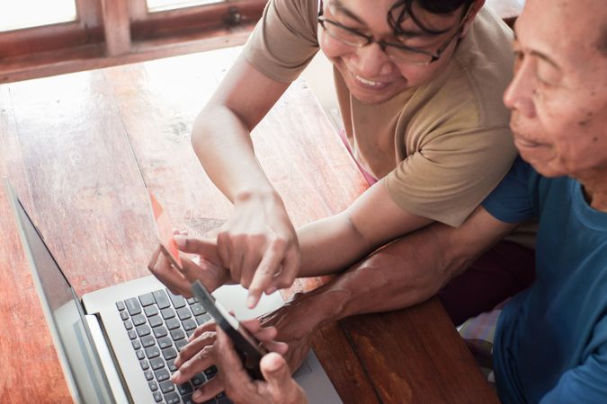 Happy adult son teaching older father using laptop and smartphone