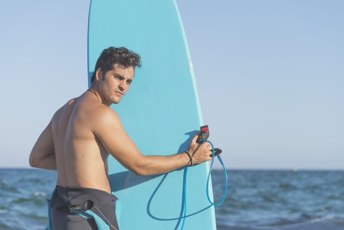Male surfer holding blue board standing in front of the sea and looking back at camerawater