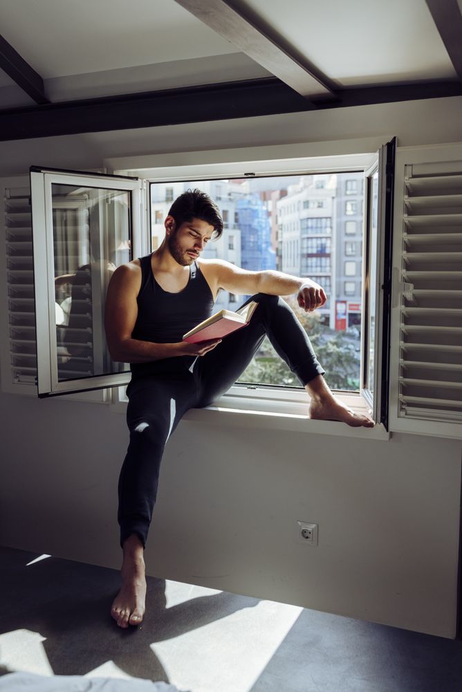 Young male sitting on window and reading book