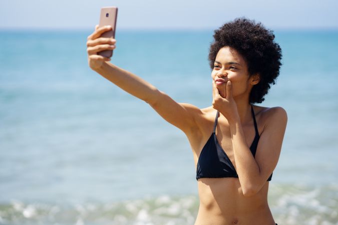 Woman in bikini taking goofy selfie on phone on coast