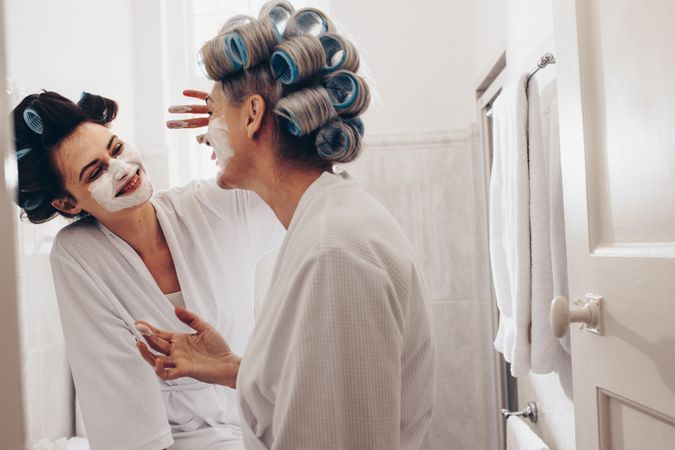 Happy adult daughter applying face cream to her mother’s face in the bathroom