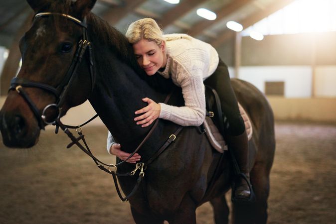 Horse owner sitting astride and lovingly hugging her brown horse