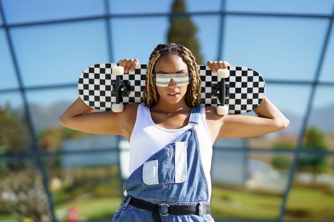 Female in denim overalls with checkered skateboard behind her shoulders