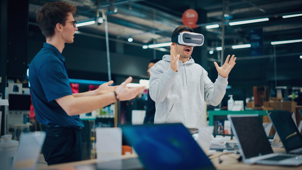 Young Man Having Conversation with a Salesperson in an Electronics Store while Trying Out a Virtual Reality Device.