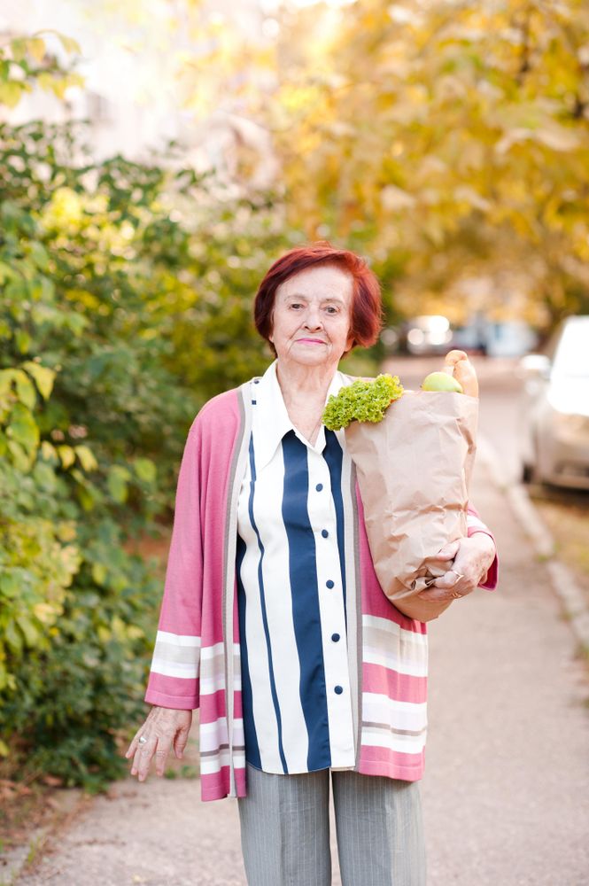 Older woman in pink cardigan holding grocery bag standing beside trees