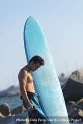 Male surfer with blue board standing in the water around a rocky beach 4dVlE0
