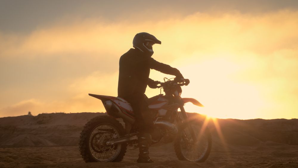 Professional FMX Motorcycle Rider Rests on His Bike and Overlooks Hard Sandy Off-Road Terrain.