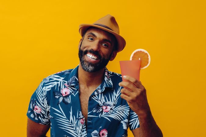 Male smiling wide with a cocktail while wearing colorful shirt with yellow background