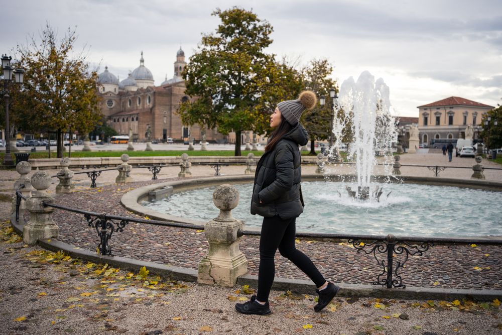 Young female tourist in warm wear strolling at ancient park with beautiful buildings and fountain