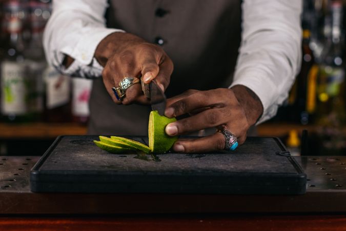 Close-up of a stylish Black bartender in a hat cutting limes to prepare cocktails