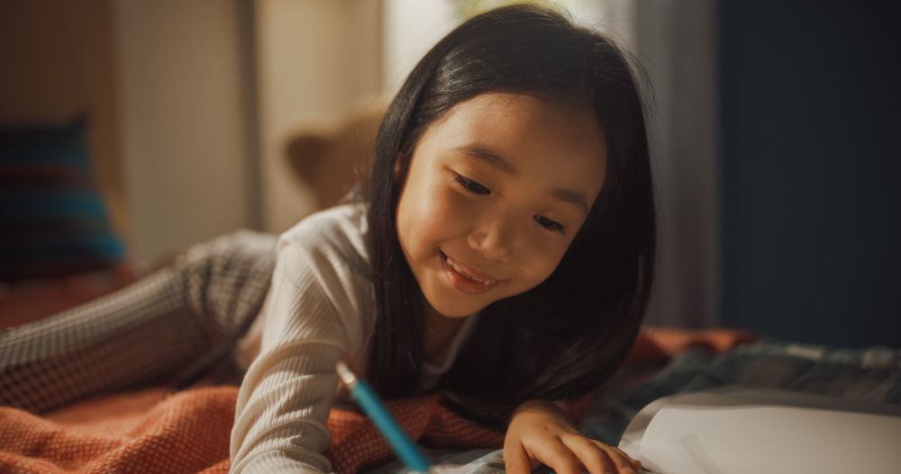 Portrait of a Little South Korean Girl Drawing with Colored Pencils in a Notebook While Lying in Bed in Children's Bedroom at Home.