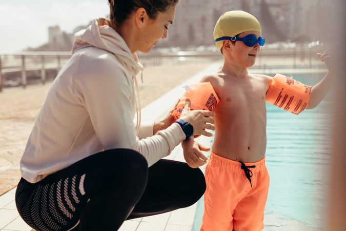 Female coach helping boy put on arm floats by poolside