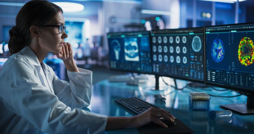 Medical Research Center Laboratory: Female Neurologist Using Desktop Computer With MRI And PET Scans Of a Brain.