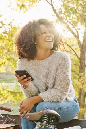 Laughing woman with curly hair using smartphone on bench in park