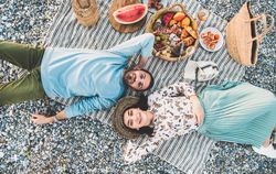 Couple lying next to picnic with watermelon, breads, oranges, and wine, looking at camera