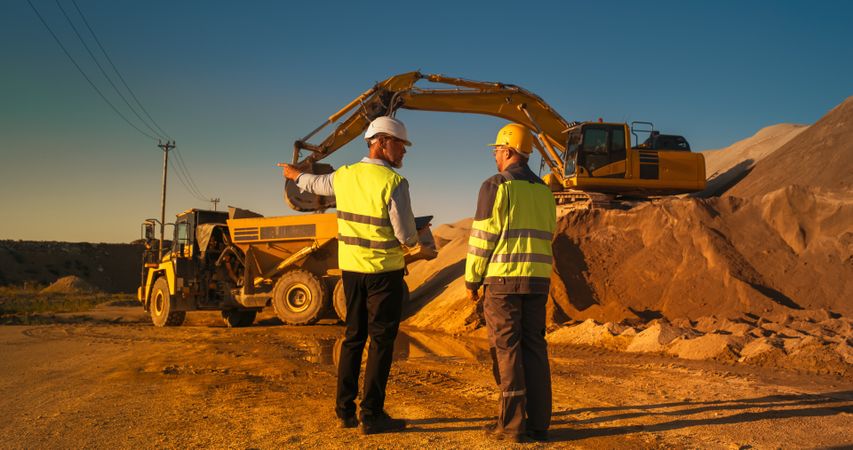 Back View Of White Real Estate Investor With Tablet And Project Manager Talking On Industrial Construction Site. Colleagues Wearing Hard Hats And Watching Heavy Machinery Working. Sunny Day