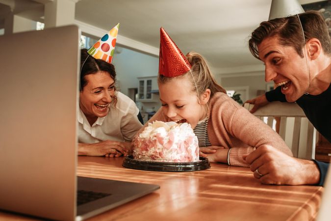 Girl biting into her birthday cake with her mouth with her parents smiling by