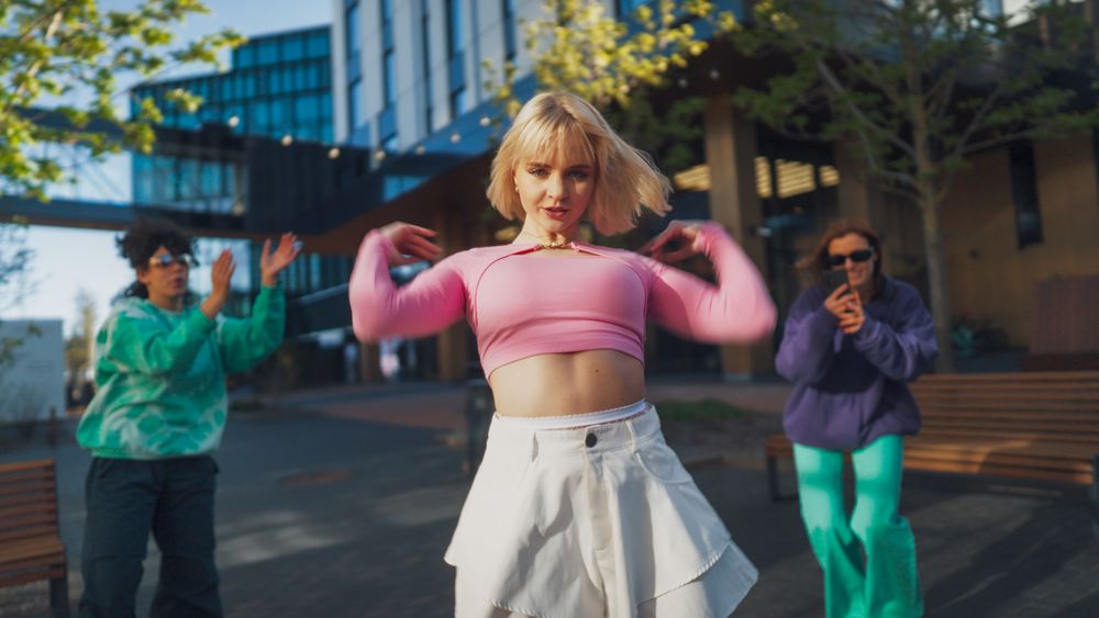 Outdoor Scene Captures A Young Blonde Woman Dancing Energetically In A Pink Top And White Skirt