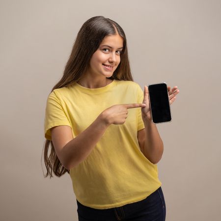 Smiling young woman pointing at a blank smartphone screen on a studio background.