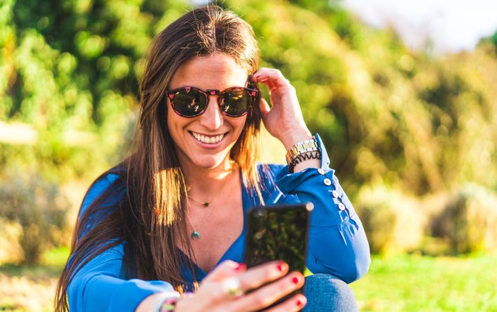 portrait of young woman with sunglasses using smartphone sitting on grass in park