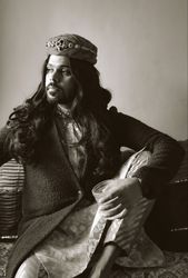 Grayscale photo of Indian man with long hair sitting on floor holding a drink