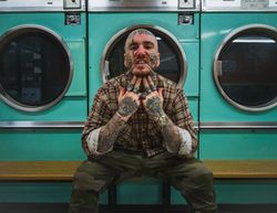 Edgy bald man with face tattoos in plaid shirt biting chain necklace in laundromat