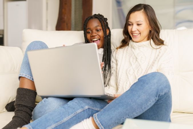 Two women in jeans and sweater watching something together on a sofa on a laptop