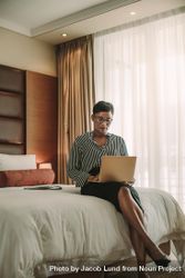 Woman sitting on bed in hotel room using laptop 4jEBx5
