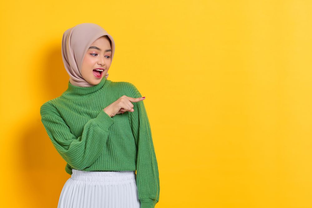 Woman in headscarf pointing to her side at yellow copy space