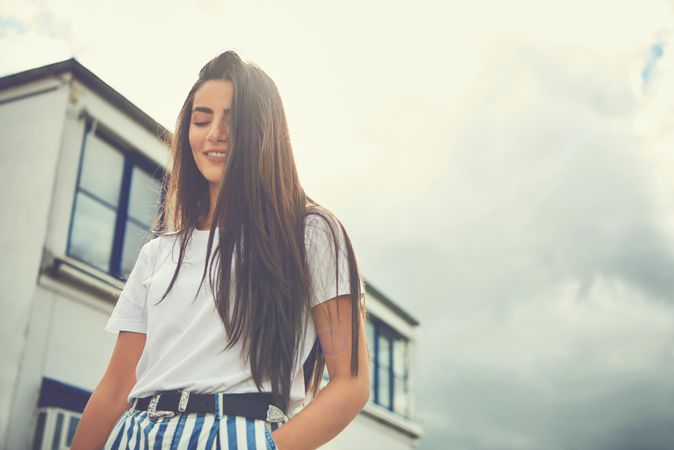 Calm woman with long brown hair looking down standing next to a building