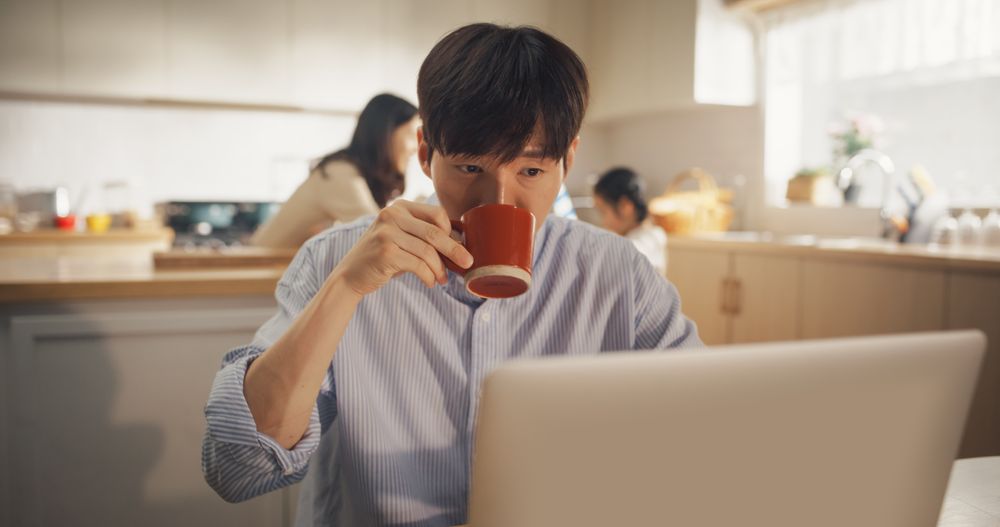 Portrait of Korean Family at Home: Young Father Using Laptop Computer for Working Remotely, With his Wife and Kids Preparing Lunch in the Background.