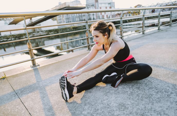 Woman with earphones and smartphone stretching on embankment