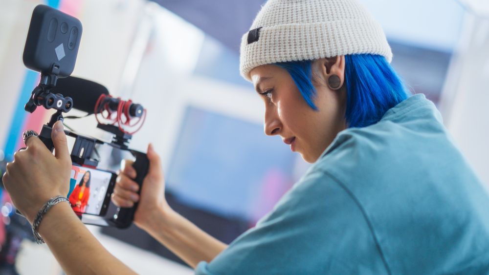 Focused Young Female Cinematographer With Blue Hair Adjusts Camera Settings On Film Set, Capturing Vibrant, Creative Video Content In A Professional Studio Environment.