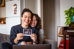 Two women smiling revealing a sonogram picture on video chat